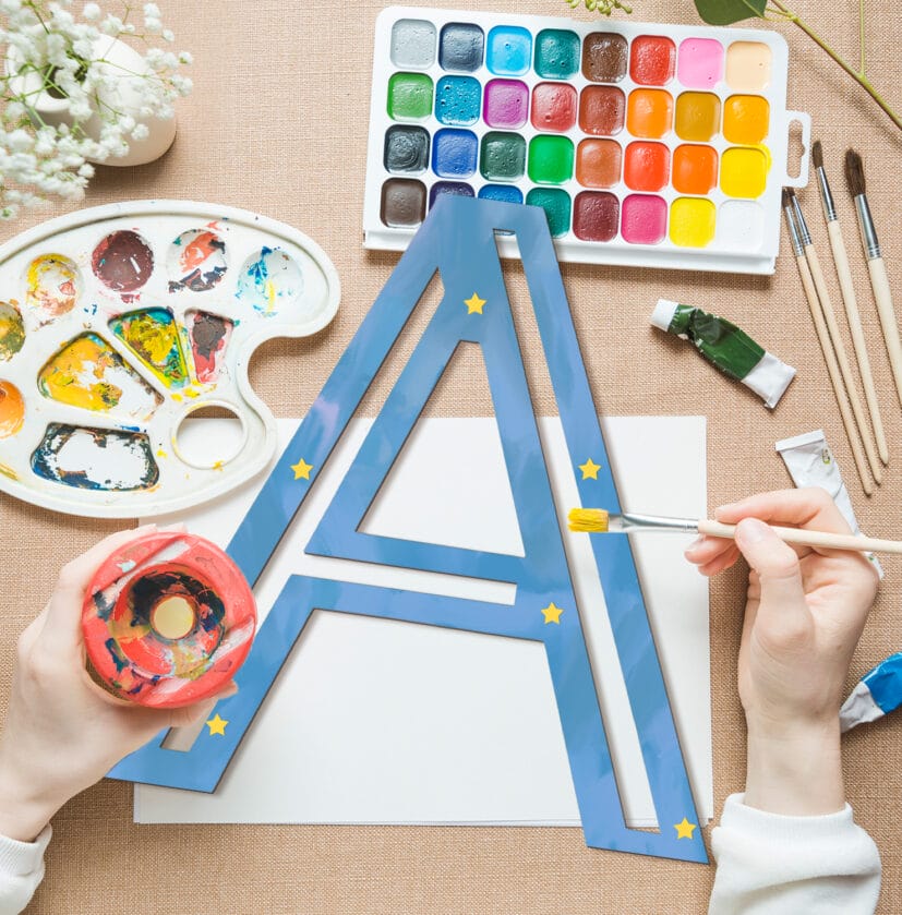 Hands paint a large blue 'A' decorated with yellow stars, surrounded by art supplies.